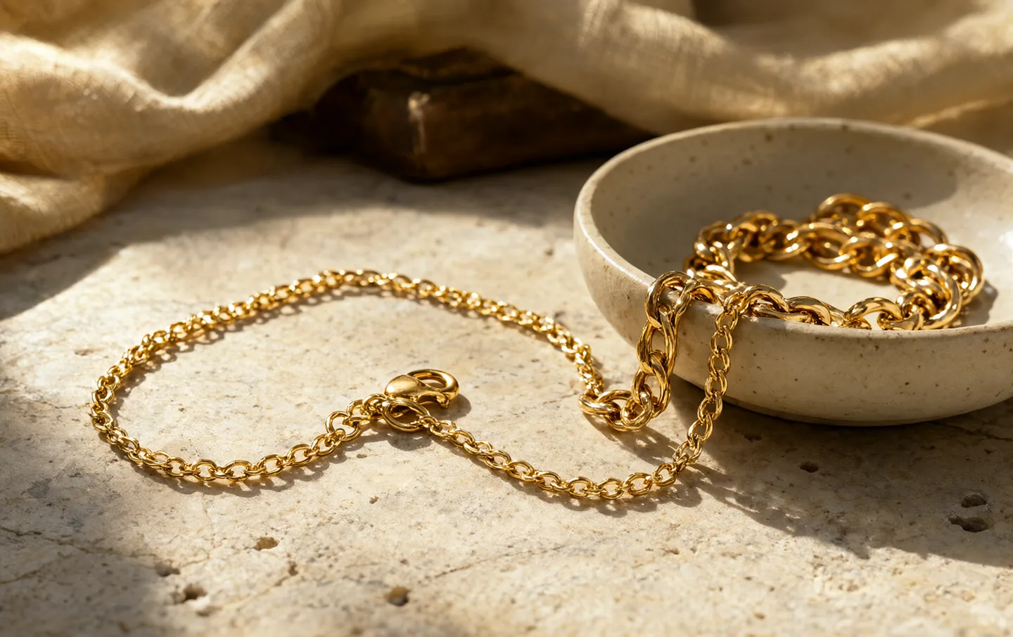 A fine gold chain coiled on a pale ceramic dish beside folded linen